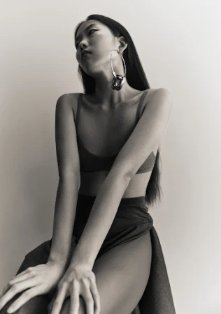 East Asian woman wearing large silver hoop earrings and a dark top seated against a plain light wall.