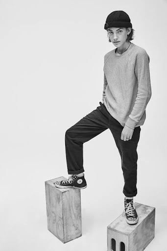 Black and white shot of a young man in beanie, sweater, and pants, standing on wooden blocks in sneakers against a plain white background.