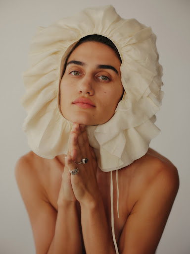 Woman wearing a white, ruffled bonnet with her hands held under her chin in a portrait against a light background.