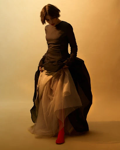 A woman in a long, layered dress poses in a studio with warm lighting. Her dark hair and the dress contrast with a light skirt and backdrop.
