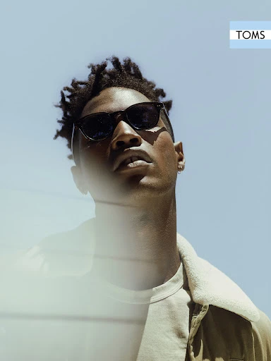 Portrait of a Black male model in sunglasses, looking up with a blue sky background and a 'TOMS' brand logo in the corner.