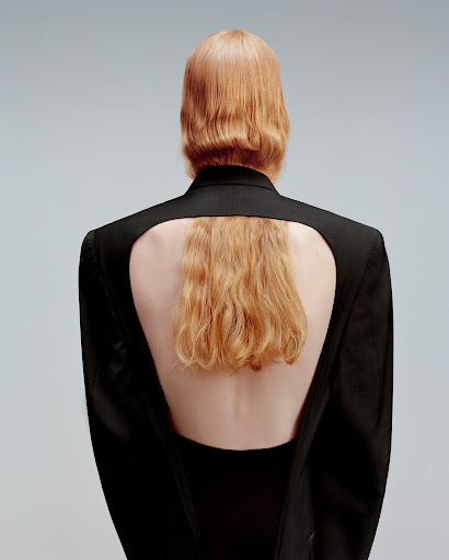 Back view of a redhead woman in a black blazer with an open back, showcasing bare skin and stylish fashion against a neutral backdrop.
