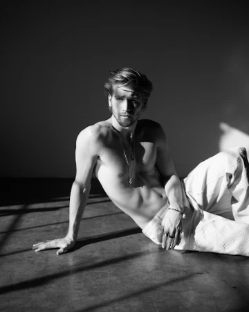 Shirtless man sitting on a studio floor wearing light trousers, illuminated by dramatic hard light and shadow patterns.