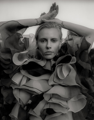 Black and white portrait of a light-skinned adult female in ruffled collar raising arms overhead, with light background out of focus.