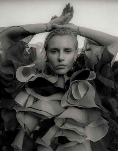 Black and white portrait of a light-skinned adult female in ruffled collar raising arms overhead, with light background out of focus.