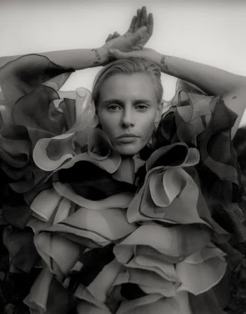 Black and white portrait of a light-skinned adult female in ruffled collar raising arms overhead, with light background out of focus.