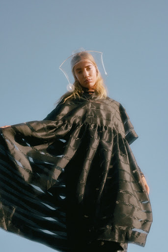 A young adult woman poses in an open outdoor setting with blue skies, wearing a dark dress and a clear plastic headpiece. 
