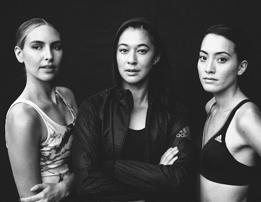 Black-and-white portrait of three female athletes in Adidas athletic wear, posing against a dark backdrop in a medium shot.