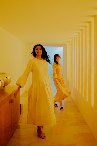 Two women in long yellow dresses walk through a bright hallway. Indoor setting, natural light, fashion editorial, minimalist composition.