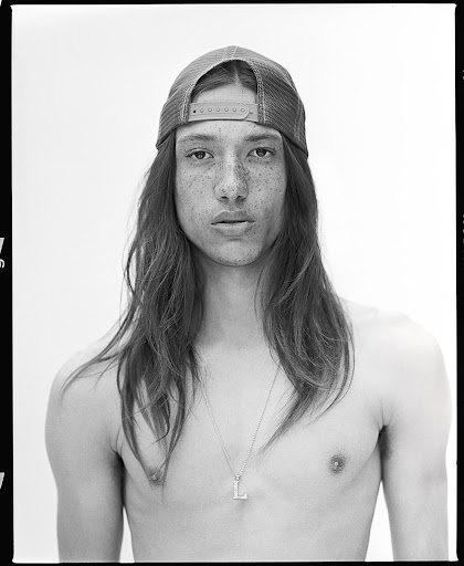 Monochrome portrait of a shirtless young man with long brown hair and a baseball cap, standing indoors against a plain white background.