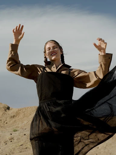 Woman in black dress and tan jacket standing in a desert landscape with arms raised against a blue sky with light clouds.