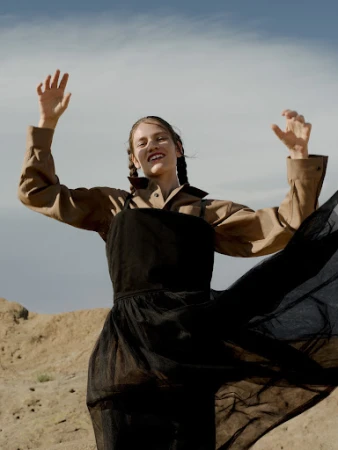 Woman in black dress and tan jacket standing in a desert landscape with arms raised against a blue sky with light clouds.