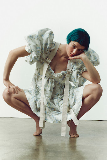A young adult woman with short teal hair poses in a white floral dress with shoulder straps against a white backdrop in a studio setting.