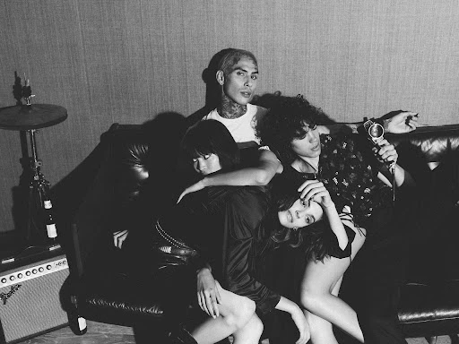 Black and white portrait of four young adults posing casually on a couch in a music studio setting with instruments in the background.
