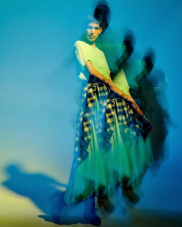 Full-length portrait of a slender person with a short haircut, wearing a yellow shirt and plaid skirt, posing in a photo studio.