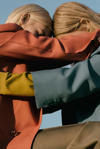 Close-up of two blond women in embracing, wearing colorful jackets in blue sky. Fashion-forward embrace in an outdoor setting.