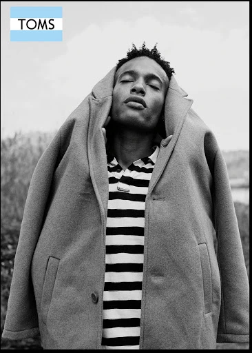 Black and white shot of a Black male model in a tan coat and striped shirt, outdoors with eyes closed. TOMS logo is visible.