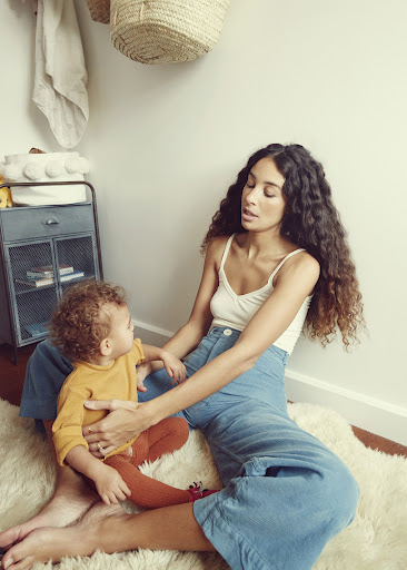 A woman with curly hair sits on a beige rug with a young child wearing a mustard-colored shirt and orange leggings. Indoor shot.