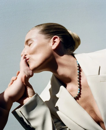Woman in an off-white blazer gently kissing an infant foot, styled by Heather Rest against a plain grey background.