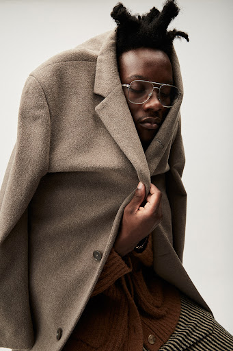 A Black man with glasses is wrapped in a tan coat, eyes closed, wearing a brown sweater and striped pants, against a white backdrop.