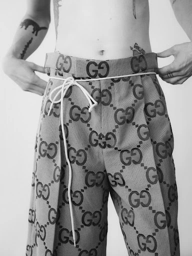 Close-up of a person adjusting the waist of patterned Gucci trousers, secured with a white rope, against a plain white backdrop.