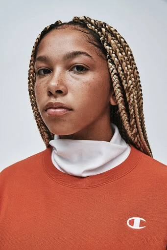 Close-up shot of a young adult female with blonde braided hair and an orange Champion sweatshirt against a plain grey backdrop.
