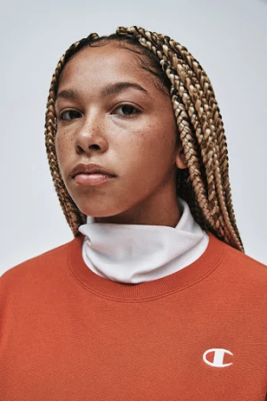 Close-up shot of a young adult female with blonde braided hair and an orange Champion sweatshirt against a plain grey backdrop.