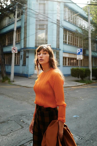 A fashionable woman in an orange sweater and plaid skirt poses on a city street near a light blue building with green trees.