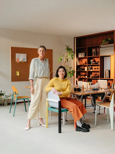 Two women pose in a modern office setting. One woman sits on a chair in orange pants, and the other stands in white pants.