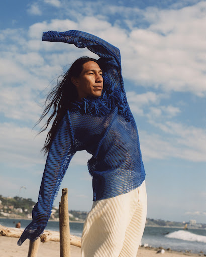 Young man poses on the beach wearing a blue mesh top and white pants against a backdrop of blue sky, clouds, and ocean waves.