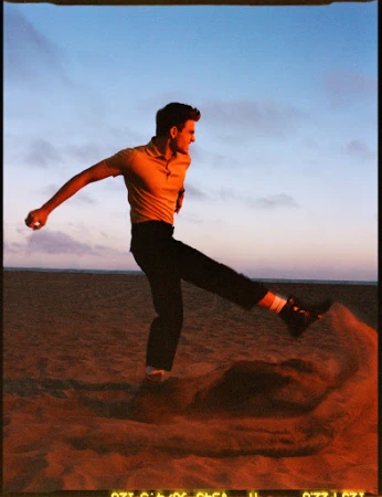Man in yellow polo shirt kicking sand on a beach at sunset styled by Heather Rest