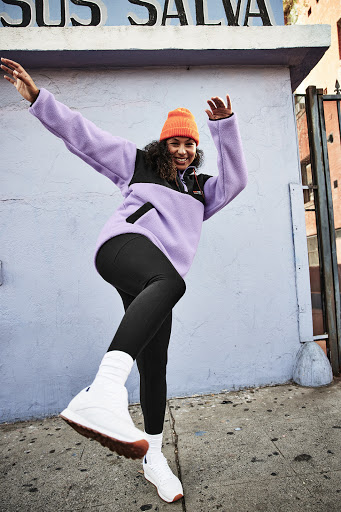 A woman poses dynamically in athletic wear on a city street, wearing an orange beanie, lavender fleece jacket, and white sneakers.