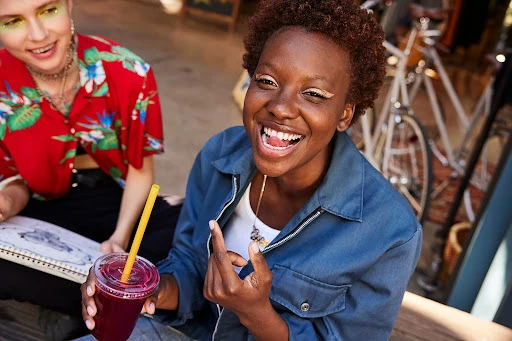 Two smiling women sitting at an outdoor cafe. One woman holds a fruit smoothie, the other is drawing. Bicycles are in the background.