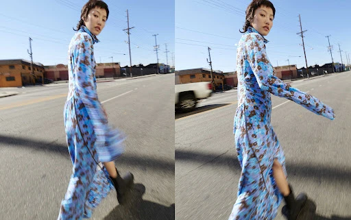 A model walking on a sunny urban street wearing a long blue patterned dress and black boots, captured with motion blur.