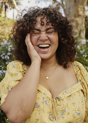 A plus-size woman with curly hair is laughing outdoors, wearing a floral print dress and gold necklaces in a natural setting.