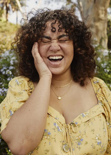 A plus-size woman with curly hair is laughing outdoors, wearing a floral print dress and gold necklaces in a natural setting.