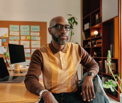 African American man in glasses poses indoors in front of laptop and corkboard with notes. Modern casual fashion, home office setting.