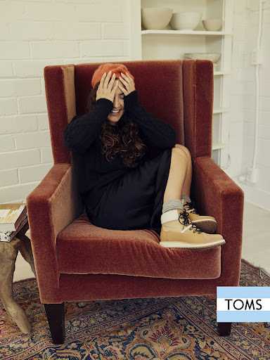 Adult woman sits in a rust-colored wingback chair indoors, wearing a beret, dark sweater and dress, with tan shoes, posing with hands on her head.