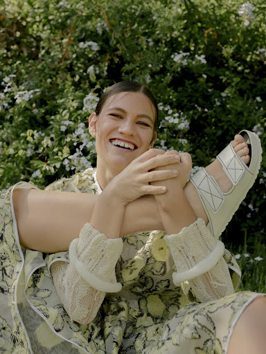 Smiling female model styled by Heather Rest wearing a patterned dress and white sandals outdoors amidst green foliage.