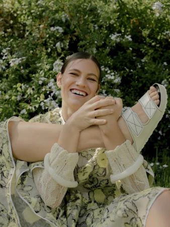 Smiling female model styled by Heather Rest wearing a patterned dress and white sandals outdoors amidst green foliage.