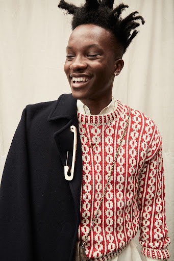 Smiling Black man in a patterned sweater and coat with a large safety pin. Stylish indoor portrait with textured background.