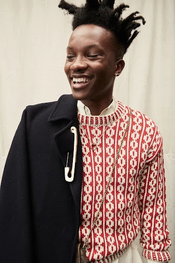 Smiling Black man in a patterned sweater and coat with a large safety pin. Stylish indoor portrait with textured background.