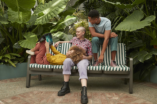 Three people relax on a bench outdoors surrounded by lush greenery. One person is reading a tablet. Casual summer clothes.