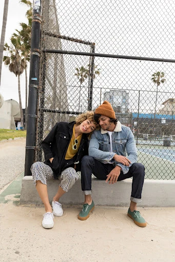 Two young adults sit together on a concrete ledge in front of a chain-link fence, posing in casual outfits near palm trees and a building.