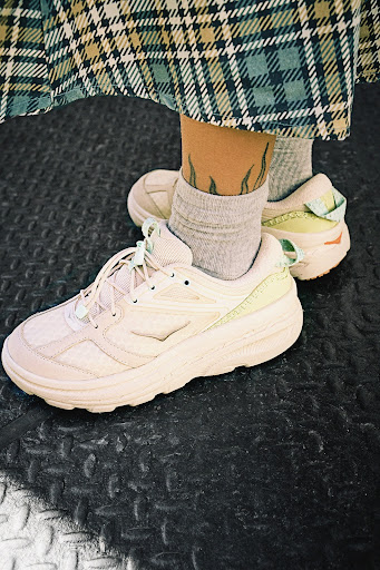 A close-up of a person wearing light-colored sneakers, grey socks, and a plaid skirt on a black textured surface.  Fashionable footwear.