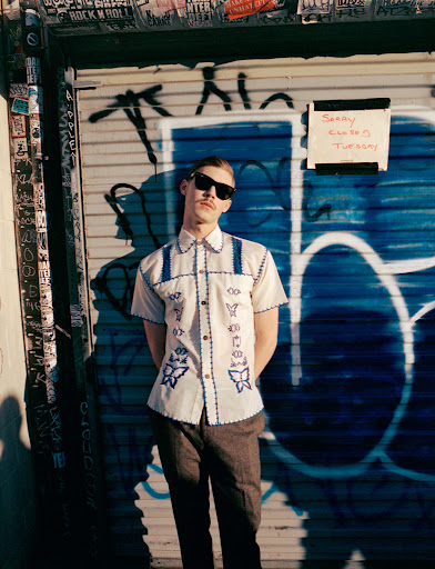 Man with mustache poses in front of metal shutter door in vintage shirt and sunglasses in the sunlight with a playful expression.