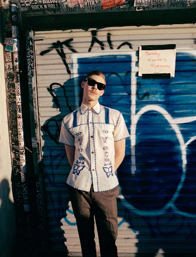 Man with mustache poses in front of metal shutter door in vintage shirt and sunglasses in the sunlight with a playful expression.