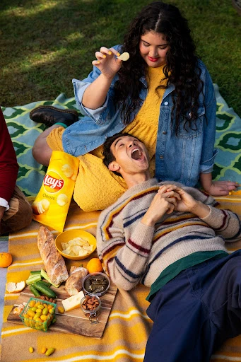 Outdoor picnic with two people; a woman holds a potato chip above a reclining man surrounded by snacks and a bag of Lay's Classic chips.