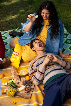 Outdoor picnic with two people; a woman holds a potato chip above a reclining man surrounded by snacks and a bag of Lay's Classic chips.