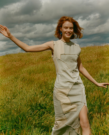 A smiling young woman with red hair stands in a tan, grassy field under a cloudy sky, wearing a patchwork dress with a side slit.